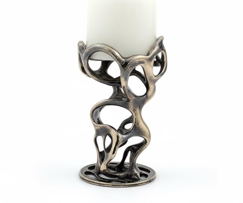 May include: An abstract bronze-colored candle holder with a white candle. The holder has an open, organic design with a circular base and a top that cradles the candle. The candle is a thick, cylindrical shape.