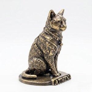 May include: A bronze-colored cat figurine in a seated position, wearing a detailed harness with a small bell. The cat is mounted on a circular base with the name ATHENA embossed in capital letters on the front edge, featuring a metallic finish.