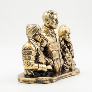 May include: A metallic gold-toned sculpture depicting three figures standing side-by-side on an oval base. The figures are dressed in winter clothing, including puffer jackets and knit beanies. The sculpture features a weathered, textured finish.
