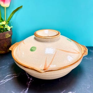 Puede incluir: Una olla de cerámica beige con tapa y borde y asas marrones. La tapa presenta puntos verdes y diseños anaranjados en forma de hojas. La olla está sobre una superficie oscura con un fondo azul.