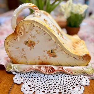 Peut inclure: Un plat de service ancien en céramique crème, avec une poignée, orné de motifs floraux jaunes et oranges. Le plat repose sur un socle décoratif à bord festonné, posé sur un napperon en crochet blanc. Des tulipes roses sont en arrière-plan.