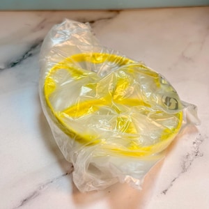 May include: A bright yellow circular plastic mold with three distinct interior compartments, encased in a clear, crinkled plastic protective bag. The mold features a smooth, rounded rim and a flat base, visible through the translucent packaging.