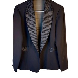 Könnte beinhalten: Ein schwarzer Blazer mit breitem Revers, das mit Reihen kleiner, dunkler, reflektierender Pailletten besetzt ist. Die Jacke verfügt über passende, mit Pailletten verzierte rechteckige Taschenklappen, lange Ärmel und ein offenes Design.