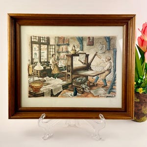 May include: A framed print depicting a vintage printing press scene. The artwork features two figures in a workshop, with a printing press, papers, and a black cat. The frame is a dark brown wood.