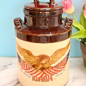 May include: A vintage ceramic milk jug with a brown lid and handle. The jug features a cream-colored base with a detailed illustration of an eagle, American flags, and the text "The Spirit of Seventy-Six". The jug has a glossy finish.