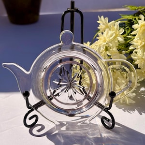 May include: A clear glass teapot-shaped trivet with a starburst design in the center, resting on a black metal stand. The trivet has a handle and spout, and is displayed on a white surface with yellow flowers in the background.