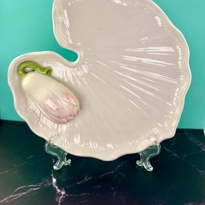 Puede incluir: Una fuente de cerámica blanca con una forma única de hoja. Presenta una flor esculpida, rosa claro y blanca, con detalles verdes. La fuente se exhibe sobre un soporte transparente, mostrando su elegante diseño. Ideal para servir aperitivos o platos pequeños.