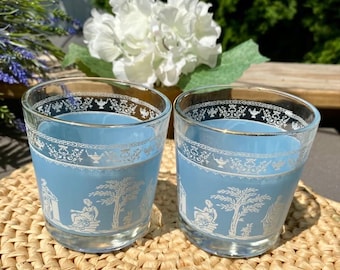Può includere: Due bicchieri vintage blu e bianchi con un design decorativo. I bicchieri presentano un interno azzurro e un motivo bianco dettagliato sul bordo e all'esterno, raffigurante figure e alberi. Sono appoggiati su un tappetino intrecciato.