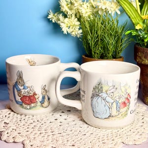 May include: Two white ceramic mugs featuring illustrations of Peter Rabbit characters. The left mug shows a rabbit in a blue coat with smaller rabbits, while the right mug depicts a rabbit in a blue dress with others. Both mugs have handles.