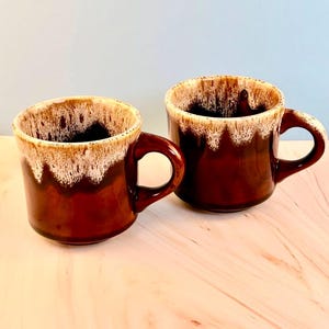 May include: Two brown ceramic mugs with a cream-colored, dripped glaze around the rim. The mugs have a rounded shape with a large handle. The mugs are sitting on a light-colored wooden surface.