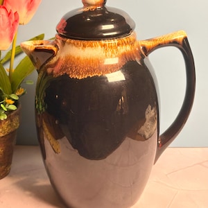 May include: A glossy ceramic coffee pot featuring a dark brown body with a lighter tan drip glaze around the upper rim and lid. The pot includes a curved handle, a small spout, and a matching removable lid with a rounded knob handle.