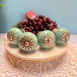 May include: Five aqua-colored cabinet knobs with silver floral designs are displayed on a white marble-like stand with gold legs. The knobs are arranged in a row, showcasing their decorative details. A pink rose and grapes are in the background.