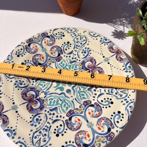 Puede incluir: Un plato decorativo de cer&aacute;mica con base crema y un patr&oacute;n detallado en azul, turquesa, rojo y negro. El plato mide aproximadamente 23 cm de di&aacute;metro. El dise&ntilde;o presenta motivos florales y arremolinados, con una regla colocada encima.