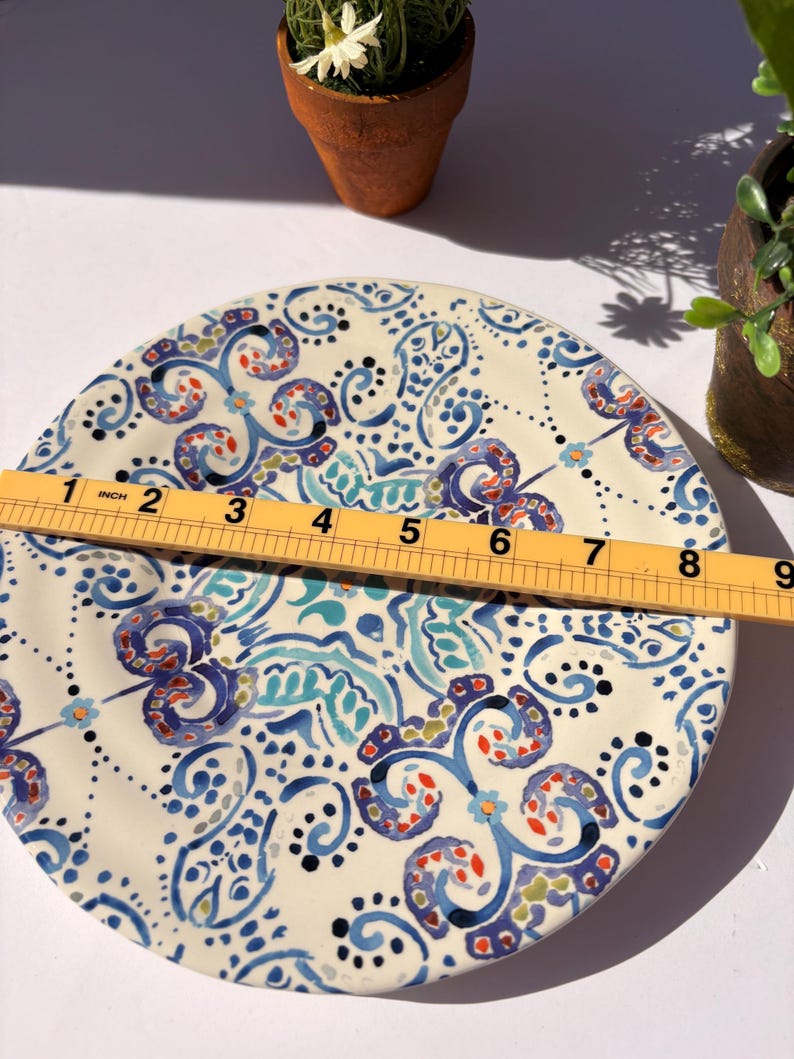 Puede incluir: Un plato decorativo de cer&aacute;mica con base crema y un patr&oacute;n detallado en azul, turquesa, rojo y negro. El plato mide aproximadamente 23 cm de di&aacute;metro. El dise&ntilde;o presenta motivos florales y arremolinados, con una regla colocada encima.