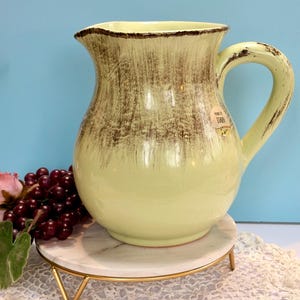 Puede incluir: Una jarra de cerámica verde claro con borde y asa marrones. La jarra tiene un acabado texturizado y envejecido y está sobre un soporte de mármol y dorado. Una etiqueta que dice "Made in Italy" está en la jarra.