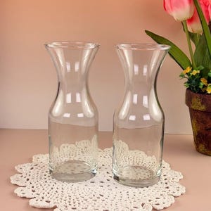 May include: Two clear glass carafes with a curved hourglass shape, sitting on a white crocheted doily. The carafes are empty and reflect the light. Pink and white tulips in a brown pot are in the background.