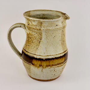 May include: A handmade ceramic pitcher with a cream-colored base and a dark brown band. The pitcher has a curved handle and a spout for pouring. The speckled design adds a rustic touch to this kitchenware.