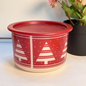 May include: A red and white Christmas-themed container with a matching lid. The container features white Christmas tree designs and white polka dots on a red background. The lid is solid red.