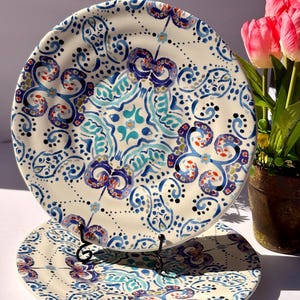 Puede incluir: Dos platos de cer&aacute;mica con intrincados dise&ntilde;os florales azules, turquesas y rojos. Los platos tienen un fondo crema y se exhiben en un soporte de metal negro. El dise&ntilde;o presenta espirales, puntos y motivos florales estilizados.