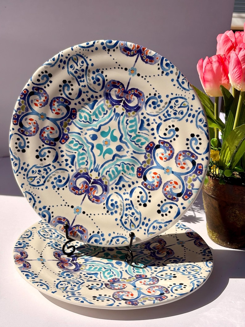 Puede incluir: Dos platos de cer&aacute;mica con intrincados dise&ntilde;os florales azules, turquesas y rojos. Los platos tienen un fondo crema y se exhiben en un soporte de metal negro. El dise&ntilde;o presenta espirales, puntos y motivos florales estilizados.