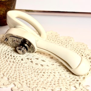 May include: A vintage off-white can opener with a curved handle and a silver cutting mechanism. The can opener is resting on a cream-colored, crocheted doily. The background includes a potted plant.