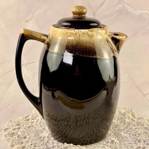 May include: A vintage ceramic coffee pot with a dark brown glaze that fades to a lighter brown at the top. The pot has a matching lid and handle, and sits on a cream-coloured lace doily.