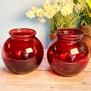 May include: Two matching spherical glass vases in a deep ruby red color. Each vase features a wide, rounded body that tapers slightly toward a smooth, circular opening at the top. The glass is translucent, allowing light to pass through the red tint.