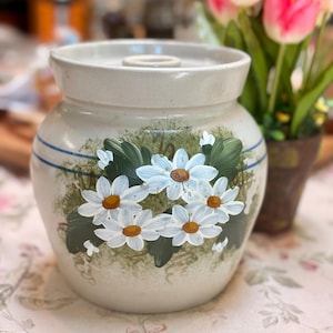 May include: A cream-colored ceramic crock with a lid, adorned with hand-painted white daisies with yellow centers and green leaves. Two blue stripes encircle the crock. A charming piece for kitchen decor.