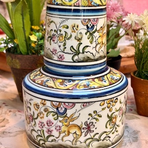 May include: A two-piece ceramic container with a lid, featuring a hand-painted design. The white base is adorned with floral and deer motifs in yellow, pink, and green, accented by blue bands. The container is approximately 6 inches tall.