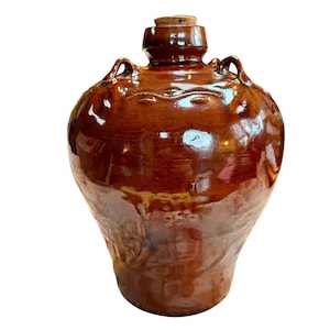 May include: A brown glazed ceramic jug with a cork stopper. The jug has a rounded body, two small handles, and decorative detailing around the neck. The jug is a rich, glossy brown colour.