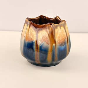 May include: A small, ceramic vase with a unique, tulip-like shape. The vase features a glossy glaze with a gradient of colors, including brown, gold, and deep blue. The rim has a wavy design, adding to its distinctive appearance.