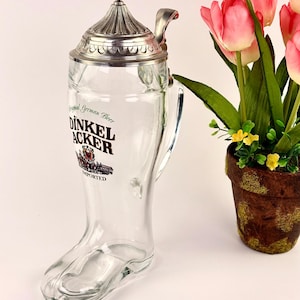 May include: Clear glass beer stein in a boot shape with a metal lid and handle. The stein has the words "DINKEL ACKER" printed on it. A bouquet of pink tulips in a brown pot is to the right of the stein.