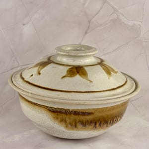 May include: A ceramic lidded bowl with a cream-colored base and lid. The bowl features brown leaf designs on the lid and a brown gradient pattern on the lower half. The lid has a small, round handle. The bowl is likely for serving or storage.