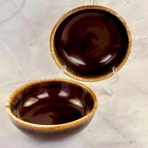 Puede incluir: Dos cuencos de cerámica marrón con acabado brillante. Los cuencos tienen un interior marrón oscuro y un borde marrón más claro. Un cuenco descansa sobre un soporte transparente, mientras que el otro está delante. Los cuencos son probablemente para servir comida.