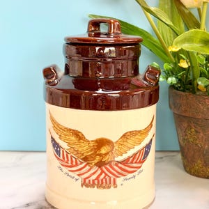 May include: A vintage ceramic jar with a brown lid and accents, featuring a cream-colored body. The jar is adorned with a patriotic design of an eagle, American flags, and the text "The Spirit of Seventy Six."