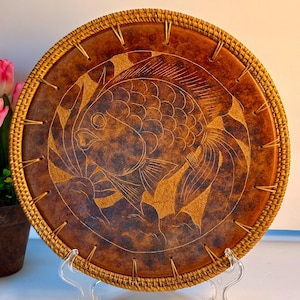 May include: Round, brown decorative plate with a fish design. The plate has a woven border and is supported by a clear stand. The fish is etched into the plate's surface, with a detailed outline.