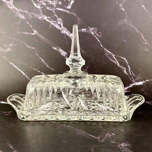 Puede incluir: Una mantequera rectangular de vidrio transparente con un adorno decorativo puntiagudo en la tapa. El plato presenta un diseño de vidrio tallado con patrones de estrellas y verticales. Tiene asas pequeñas.