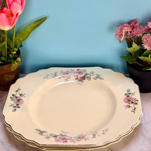 May include: A stack of three off-white, scalloped-edge plates with a floral design featuring pink flowers and green leaves. The plates have a gold trim. The plates are on a marble surface with pink tulips and pink flowers in the background.