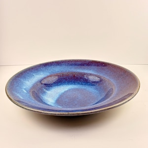 May include: A round, ceramic bowl with a deep center and a wide rim. The bowl has a glossy, iridescent finish with shades of blue and purple. The rim has a dark, metallic edge. This decorative bowl is suitable for serving food.