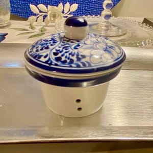Puede incluir: Un recipiente de cerámica blanca con una tapa con estampado floral azul y blanco. La tapa tiene un pequeño pomo redondo azul. El recipiente tiene dos pequeños agujeros cerca de la base. El recipiente está sobre una bandeja plateada.