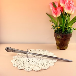 Puede incluir: Un afilador de cuchillos plateado con un mango ornamentado descansa sobre un tapete de ganchillo color crema. El afilador está colocado en diagonal, con una guarda cerca del centro. Un tiesto con tulipanes rosas está en el fondo.