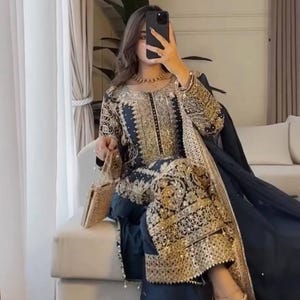 Puede incluir: Conjunto de ropa formal bordada en azul marino y dorado. El atuendo incluye una parte superior de manga larga, pantalones a juego y una bufanda fluida. Un pequeño clutch dorado y tacones dorados con correas completan el look. El atuendo está muy adornado con bordados dorados.