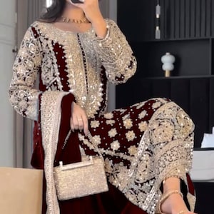 May include: A formal maroon velvet outfit with intricate gold embroidery and embellishments. The ensemble includes a long-sleeved top, matching pants, and a scarf. A small, gold, sequined handbag and gold heels complete the look.
