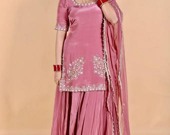 Può includere: Un tradizionale completo indiano color rosa polveroso. L'outfit include un top a maniche corte con ricami intricati e una gonna abbinata. Una sciarpa trasparente con finiture dorate è drappeggiata sulla spalla. L'ensemble è impreziosito da braccialetti e orecchini rossi.