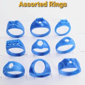 May include: A collection of nine blue wax ring models arranged in a three-by-three grid, each featuring distinct settings for gemstones. The text Assorted Rings is displayed in yellow at the top of the image, indicating a variety of ring designs.