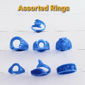 May include: A collection of assorted blue rings, each with a unique design, displayed on individual ring holders. The rings include various shapes, such as a dolphin, a lion's head, and a ring with a decorative pattern. The text "Assorted Rings" is displayed at the top.