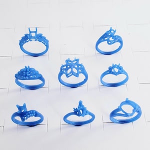 May include: A collection of eight blue wax ring castings arranged in three rows. Each ring features a distinct setting design, including floral, prong, and bypass styles, intended for jewelry making. The wax models are smooth and uniform in color.