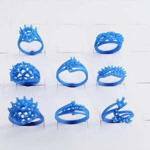 May include: A collection of eight blue wax ring models arranged in a grid. Each ring features intricate prongs and settings designed for jewelry casting, showcasing various styles including solitaire, multi-stone, and bypass band configurations.