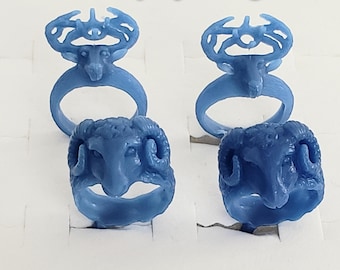 May include: Four blue trophy rings are displayed, two with deer head designs and two with ram head designs. The rings are set against a white background. The text "Trophy Rings" is displayed in yellow at the top of the image.