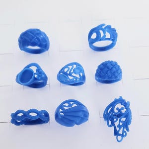 May include: A collection of eight blue wax ring models arranged in a grid. The rings feature various designs, including woven textures, openwork filigree patterns, floral motifs, and smooth geometric shapes, all cast in a solid blue wax material.